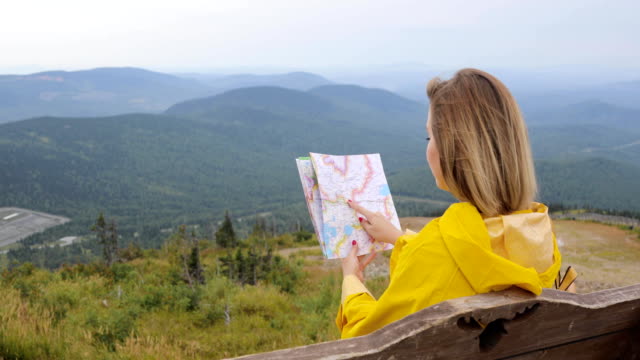 Young-female-hiking-im-yellow-raincoat-with-a-backpack-in-mountains-holding-paper-map-in-hands