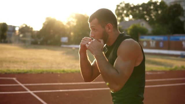 Bearded,-concentrated-man-boxer-doing-boxing-exercise,-warming-up-while-standing-on-stadium-outdoors-in-summer-sun-rays.-Side-view