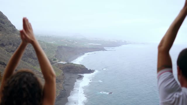 Un-hombre-y-una-mujer-sentada-en-la-cima-de-una-montaña-mirando-el-mar-sentado-en-una-piedra-de-meditación-levantando-sus-manos-y-realizando-una-respiración-relajante.-Islas-Canarias