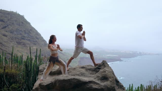 Ein-Mann-und-eine-Frau-stehen-am-Rand-einer-Klippe-mit-Blick-auf-den-Ozean-erheben-ihre-Hände-oben-und-atmen-Sie-die-frische-Seeluft-während-yoga