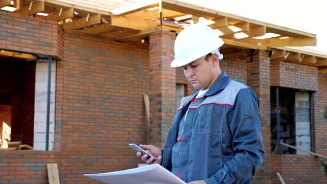 Man-in-white-hardhat-and-overall-holding-paper-plans-of-building-and-using-phone-outside-on-site