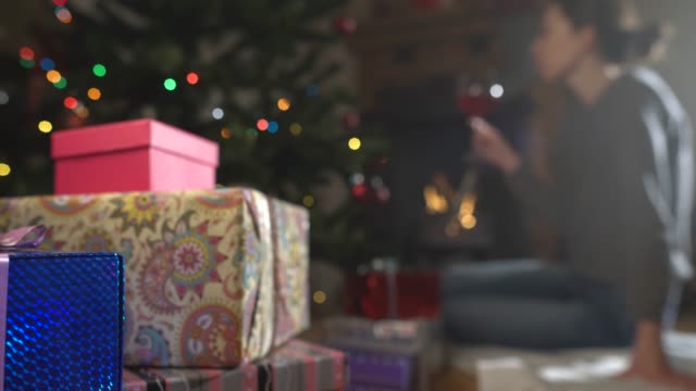 junge-Frau-trinkt-Wein-dem-geschmückten-Weihnachtsbaum-und-am-Kamin.-Geschenke-im-Fokus
