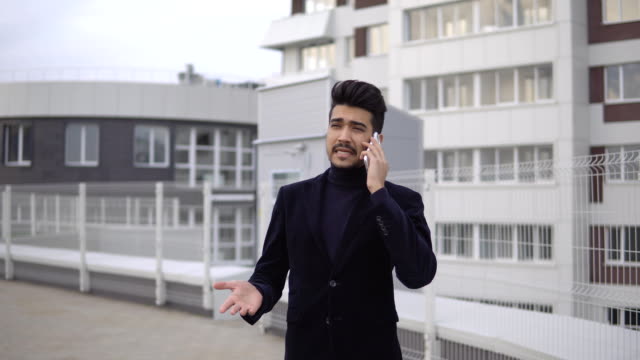 Handsome-young-business-man-talking-on-smartphone-and-walking-away-smiling-happy-wearing-suit-jacket-outdoors