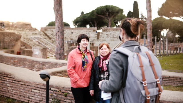 Joven-hermosa-chica-Europea-tomando-una-foto-de-dos-mujeres-mayores-cerca-de-antiguas-ruinas-históricas-en-Ostia,-Italia-de-vacaciones.