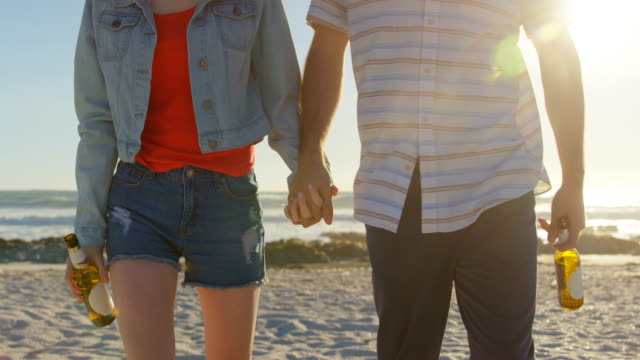 Mid-section-of-couple-holding-hands-with-beer-bottle-walking-on-beach-4k
