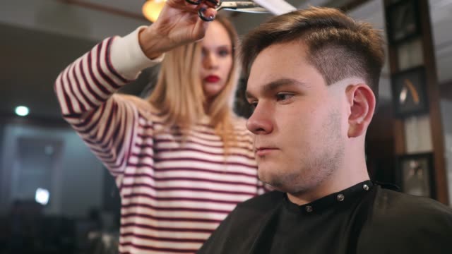 Interior-shot-of-working-process-in-modern-barbershop.-Side-view-portrait-of-attractive-young-man-getting-trendy-haircut.-Male-hairdresser-serving-client,-making-haircut-using-metal-scissors-and-comb