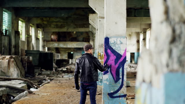 Männliche-Graffiti-Maler-schafft-abstraktes-Bild-mit-Sprühfarbe-in-verlassenen-Gebäude-leer.-Alte-Spalte-ist-im-Vordergrund,-schmutzige-Wände-und-Fenster-im-Hintergrund.
