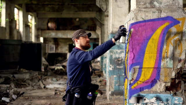 Junger-Mann-arbeiten-professionelle-Graffiti-Maler-in-verlassenen-Gebäude,-malt-er-mit-Aerosol-Sprühfarbe-auf-beschädigte-Spalte.-Moderne-Kunst-und-Kreativität-Konzept.