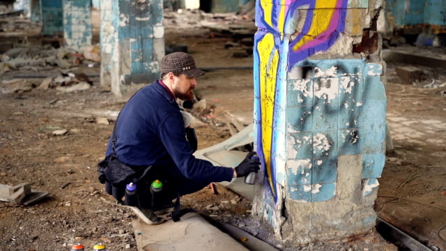 Graffiti-artist-bearded-guy-is-painting-on-pillar-in-abandoned-building-with-aerosol-paint-spray.-Empty-industrial-building-with-dirty-walls-and-floor-is-in-background.