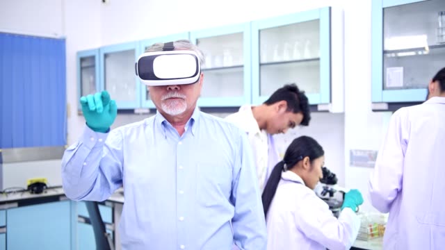 Scientists-working-in-lab-portrait.-Senior-chinese-male-scientist-working-in-lab-with-VR-head-set-pressing-the-button-portrait-with-his-team-in-background.-For-special-effect-overlay.