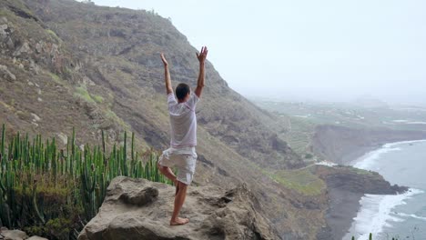 A-man-standing-on-the-edge-of-a-cliff-overlooking-the-ocean-raises-his-hands-up-and-inhales-the-sea-air-during-yoga