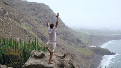 Ein-Mann-steht-am-Rand-einer-Klippe-mit-Blick-auf-den-Ozean-erhebt-seine-Hände-und-atmet-die-Luft-während-yoga