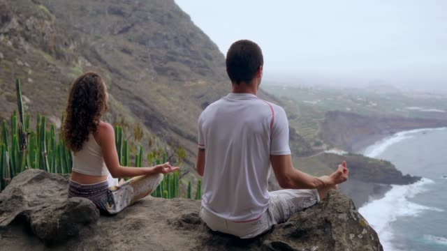 Ein-Mann-und-eine-Frau-sitzen-auf-einem-Berg,-der-Blick-auf-den-Ozean,-sitzt-auf-einem-Stein-in-einem-Lotussitz-meditieren.-Die-Ansicht-von-hinten.-Kanarische-Inseln