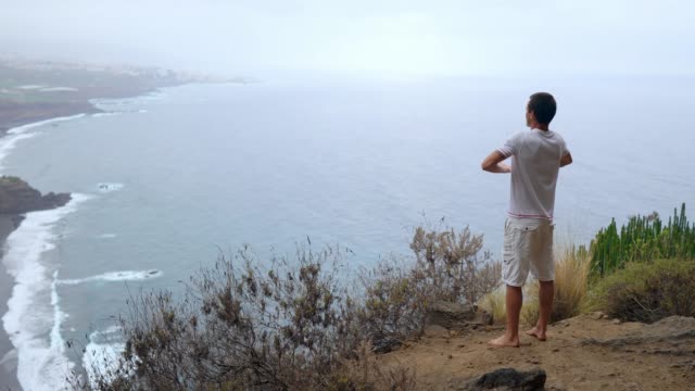 Ein-Mann-steht-am-Rand-einer-Klippe-mit-Blick-auf-den-Ozean-erhebt-seine-Hände-und-atmet-die-Luft-während-yoga