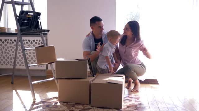 Feliz-familia-con-hijo-pequeño-disfruta-de-nueva-luz-hogar-después-de-las-reparaciones-entre-cajas-de-cartón-con-cosas