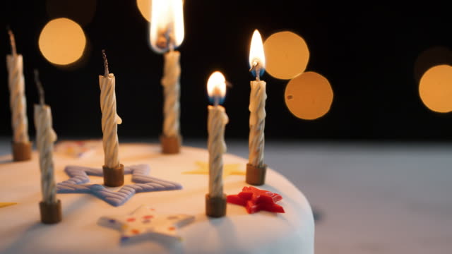 Close-up-of-four-candles-being-lit-with-a-match,-amongst-unlit-candles-on-a-white,-decorated-birthday-cake,-bokeh-lights-in-the-background