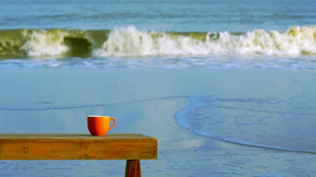 Orange-cup-of-coffee-on-brown-wooden-table-on-beach-with-surf-of-blue-sea-in-background.
