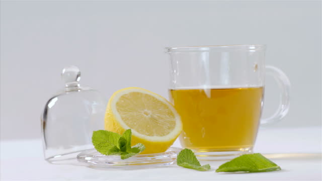 Herbal-tea-being-poured-into-transparent-teapot,-lemon-and-mint-leaves-around,-75-fps-slow-motion