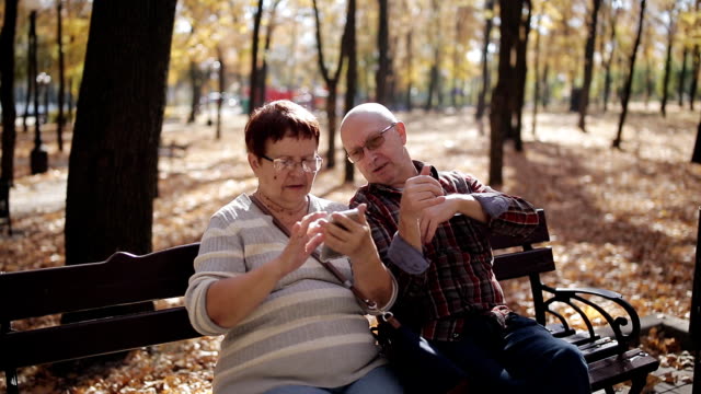 Ein-älteres-Ehepaar-im-Herbst-Park-versiert-mit-dem-Smartphone,-Fotos-durchsuchen-und-im-Internet-surfen.