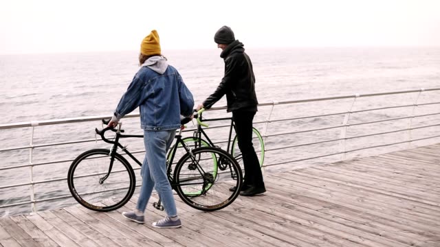 Schöne-Hipster-paar-verlassen-ihre-Fahrräder-stützte-sich-auf-Querbalken-gehen-zusammen-in-der-Nähe-des-Meeres-am-Herbsttag-umarmen.-Fuß-durch-Holzdeck-tagsüber