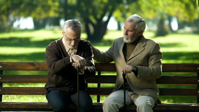 Old-man-supporting-depressed-friend,-sitting-in-park-near-nursing-home,-lost