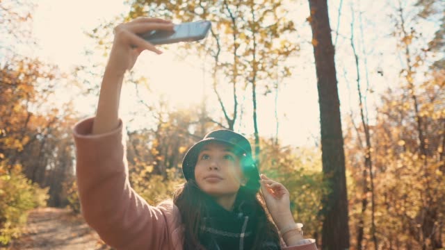 Chica-encantadora-está-tomando-selfie-por-teléfono-móvil-en-el-bosque-del-otoño-soleado,-sonriendo
