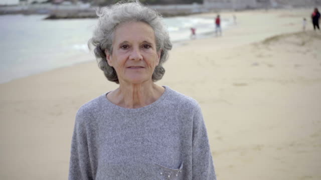 Smiling-senior-woman-walking-on-sandy-beach-and-talking-to-camera.