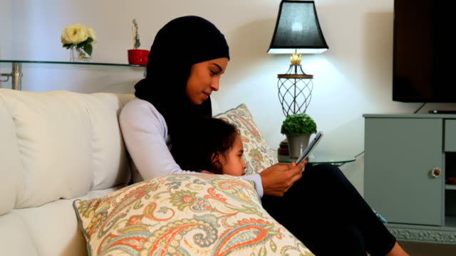 Madre-joven-usando-una-tableta-digital-con-su-hija-en-el-sofá-en-casa-4k