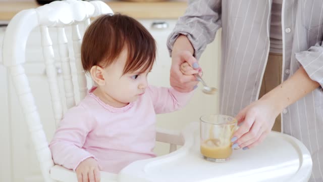 Unrecognizable-mother-helping-her-baby-daughter-eat-fruit-puree-with-spoon-in-highchair