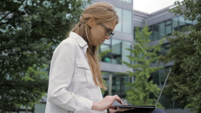 Joven-mujer-de-negocios-que-usa-computadora-portátil-en-green-park-junto-al-edificio-de-oficinas