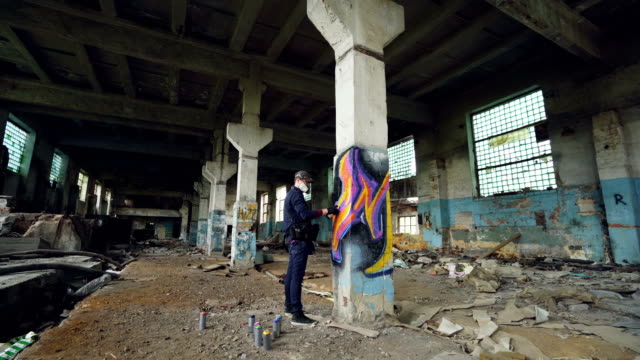 Graffiti-artist-in-protective-mask-is-painting-on-high-column-in-abandoned-industrial-building.-Creative-people,-modern-wall-art-and-protective-equipment-concept.