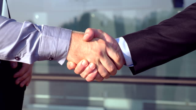 Close-up-of-successful-businessmen-greeting-each-other-near-office-building.-Two-young-colleagues-meeting-and-shaking-hands-in-urban-environment.-Handshake-of-business-partners-outdoor.-Side-view