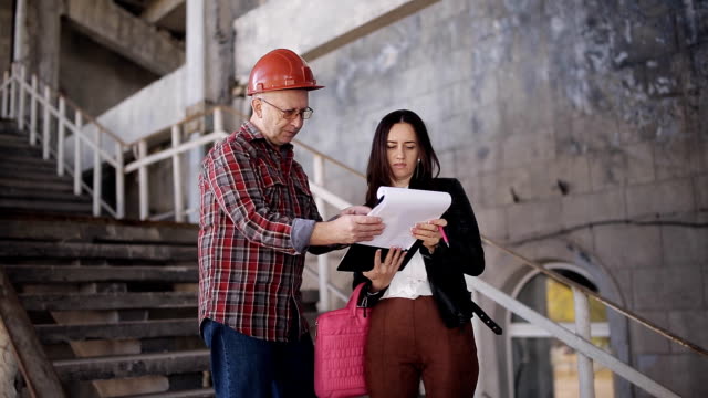 The-woman-engineer-and-the-foreman-in-helmet-compare-statement-acquired-for-construction-materials-and-confirm-the-date-of-delivery-of-the-object.