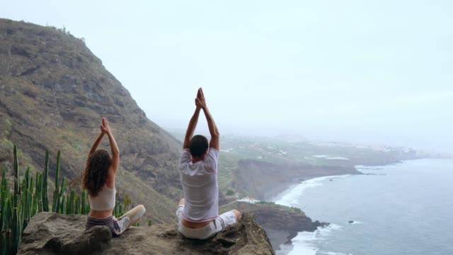 Un-hombre-y-una-mujer-sentada-en-la-cima-de-una-montaña-mirando-el-mar-sentado-en-una-piedra-meditando-en-posición-de-loto.-La-vista-desde-la-parte-posterior.-Islas-Canarias