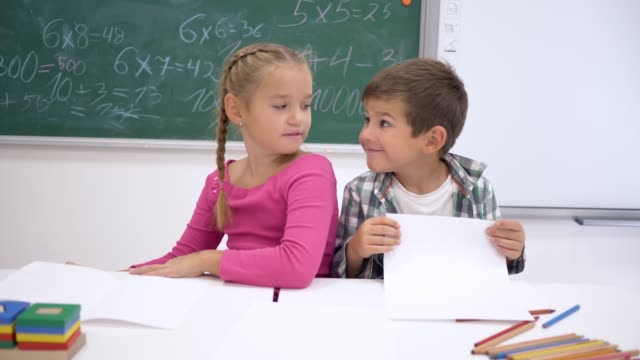 Klassenkameraden-Kinder-umarmen-und-lächelnd-sitzen-am-Schreibtisch-vor-dem-Hintergrund-der-Tafel-im-Klassenzimmer-in-der-Schule