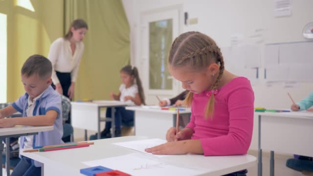 kleines-Schulmädchen-sitzt-hinter-der-Schulbank-während-Zeichnung-Lektion-in-helle-Klassenzimmer-auf-Hintergrund-der-Mitschüler-und-eine-junge-Lehrerin