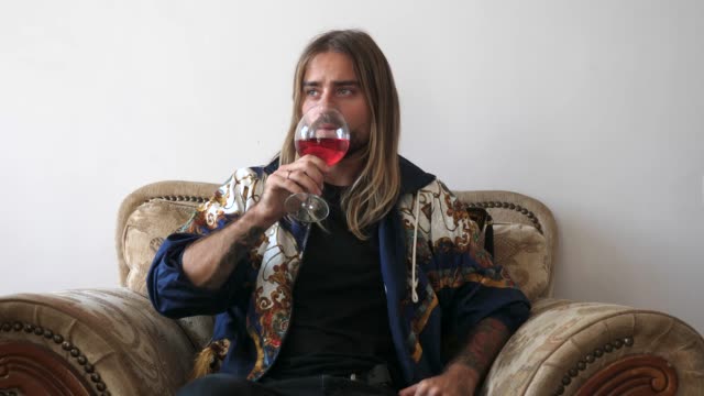 Happy-young-man-sitting-on-a-comfortable-armchair-with-cushions-wearing-a-jacket-inside-his-home-holding-a-glass-of-delicious-red-wine-tasting-and-swirling