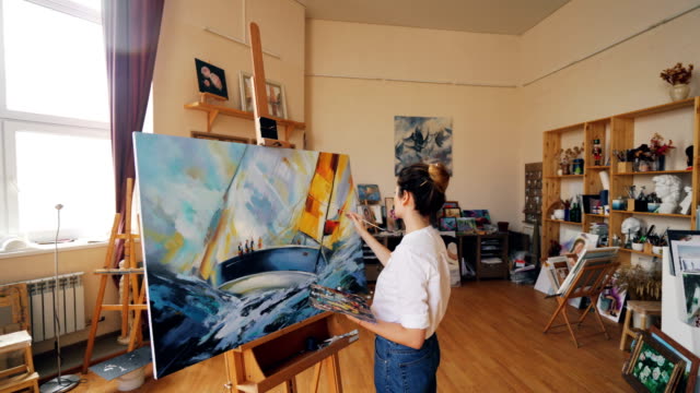 Pan-shot-of-female-painter-depicting-marine-landscape-on-canvas-using-oil-paints,-brush-and-palette-working-in-workshop-alone.-Visual-arts-and-youth-concept.
