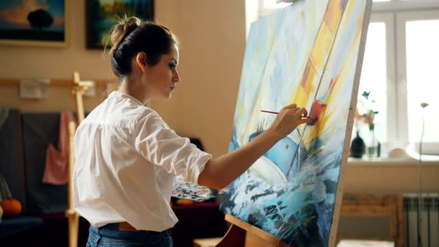 Professional-artist-young-woman-is-painting-seascape-with-acrylic-paints-finishing-marine-landscape-ship-and-sea-waves-working-alone-in-cozy-studio.-People-and-work-concept.