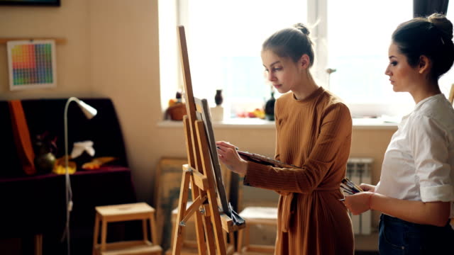 Cheerful-art-student-is-working-at-picture-painting-with-brush-on-canvas-while-her-teacher-is-checking-her-work-and-helping-her.-Girl-is-smiling-enjoying-class.
