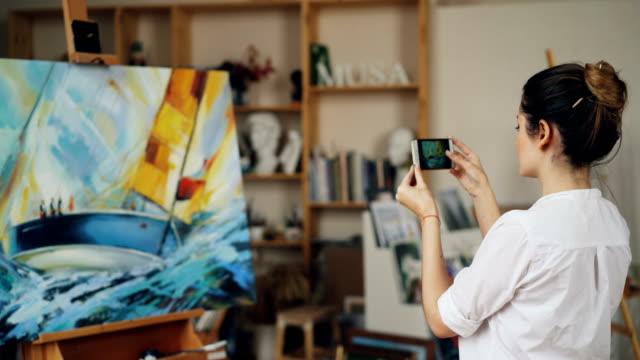 Malerin-nimmt-ein-Foto-von-ihr-schönes-Bild-mit-modernen-Smartphone-Kamera-Bildschirm-stehen-im-Studio-allein-zu-berühren.-Moderne-Technik-und-Kunst-Konzept.