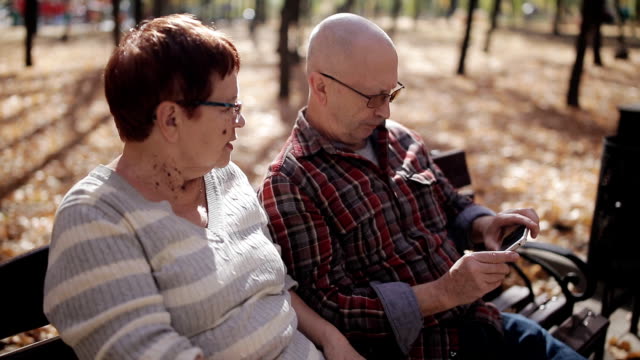 Ein-älteres-Ehepaar-im-Herbst-Park-auf-einer-Bank,-die-Bilder-von-mir-auf-dem-Smartphone.