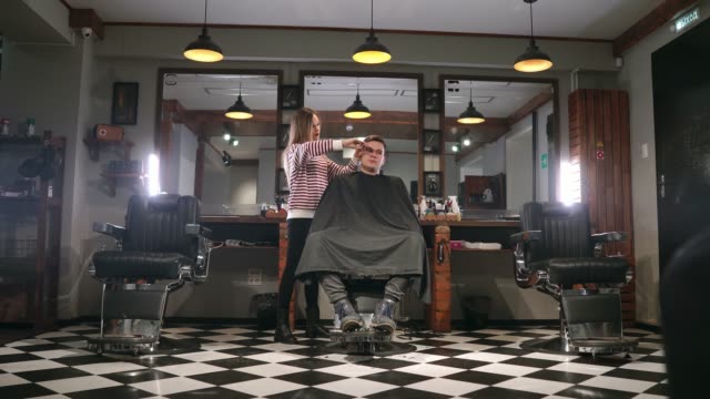 Interior-shot-of-working-process-in-modern-barbershop.-Side-view-portrait-of-attractive-young-man-getting-trendy-haircut.-Male-hairdresser-serving-client,-making-haircut-using-metal-scissors-and-comb