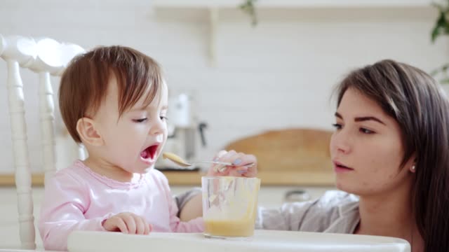 Close-up-side-view-of-young-mother-feeding-her-baby-daughter-vegetable-puree-with-spoon,-girl-trying-to-feed-herself