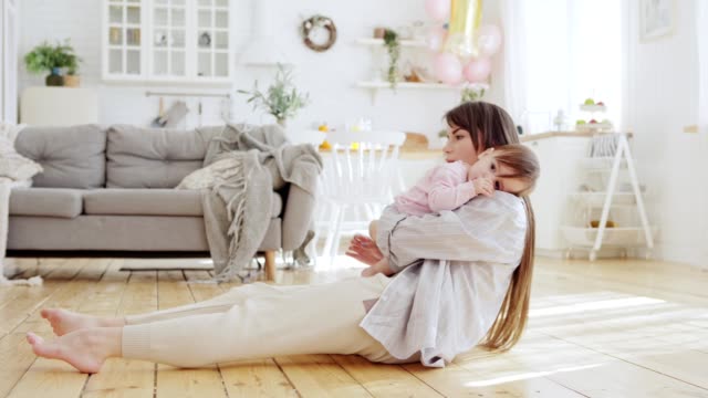 Young-mother-lying-on-hardwood-floor-in-domestic-room-embracing-her-cute-baby-daughter