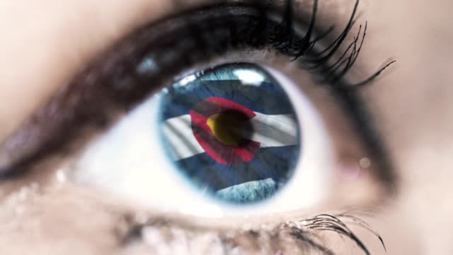 Woman-blue-eye-in-close-up-with-the-flag-of-Colorado-state-in-iris,-united-states-of-america-with-wind-motion.-video-concept