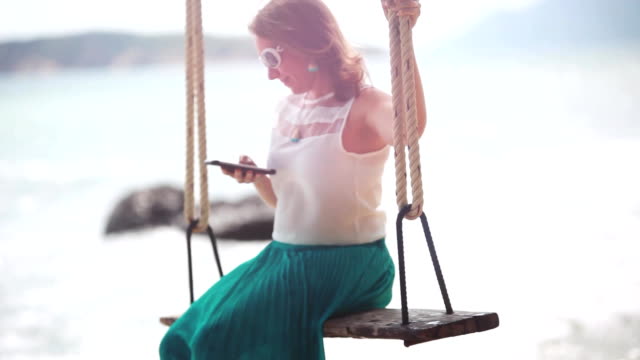 Young-happy-woman-wearing-sunglasses-uses-mobile-phone-on-a-swing-on-a-tropical-beach-and-splashes-waves-beautiful-breaks-by-stones-in-the-sea.-1920x1080