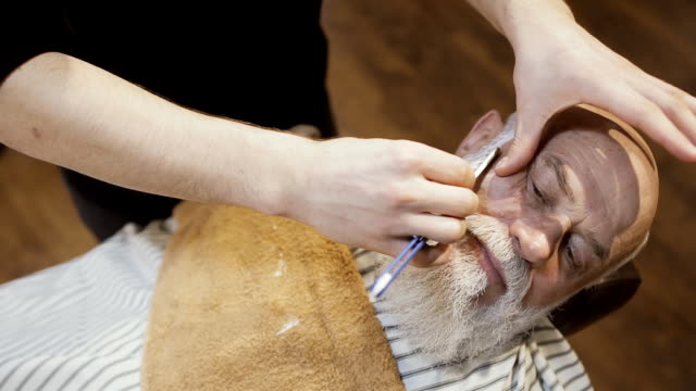 Barbier-rasiert-Bart-des-älteren-Mannes-mit-Klinge-in-barbershop