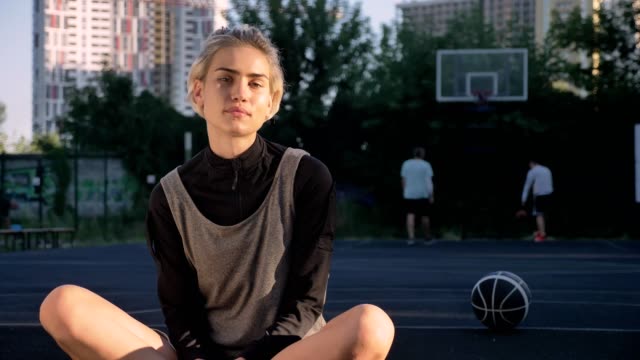 Gorgeous-blonde-woman-sitting-on-basketball-court-and-smiling-at-camera,-men-in-background-playing,-female-player