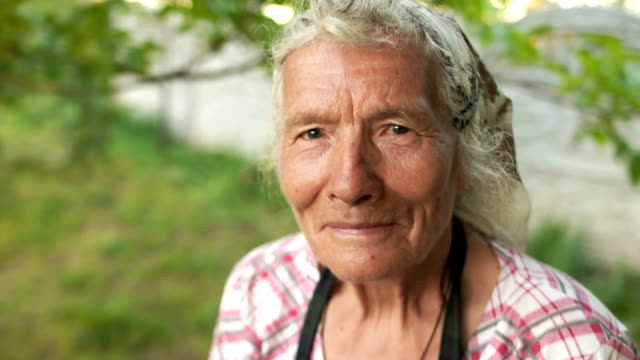 Portrait-of-an-elderly-woman-in-a-kerchief.-The-wind-wavers-her-gray-hair,-she-looks-into-the-camera-and-blinks.-Grandmother-in-the-garden-near-the-house.-Close-up